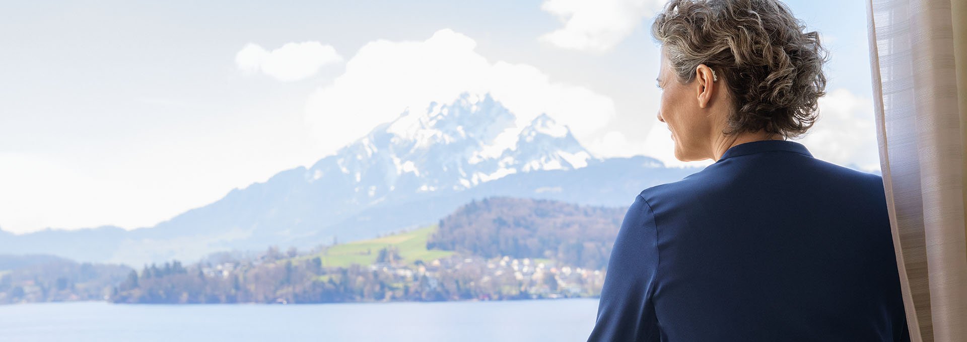 Woman wearing Bernafon Alpha rechargeable hearing aids enjoys Swiss lake and mountain view from hotel window