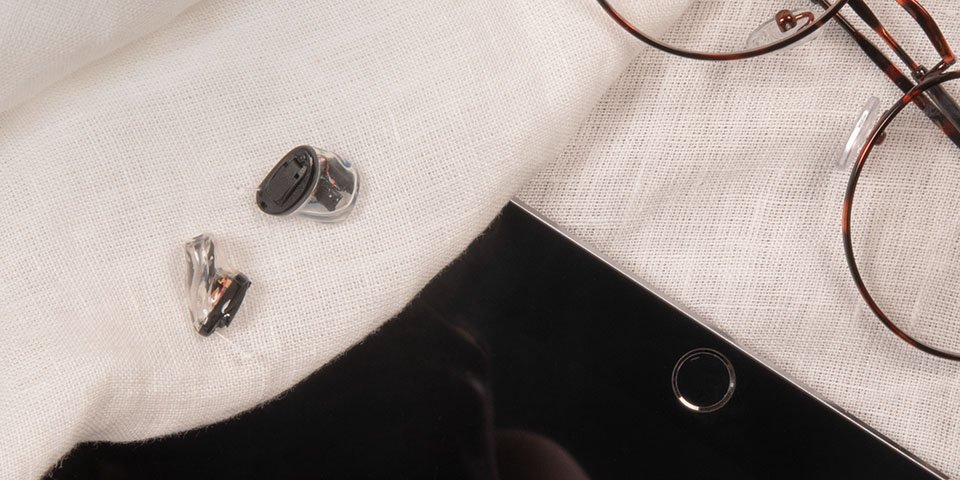 The black and transparent smallest in the ear hearing aids lying on a white sheet next to eyeglasses and tablet.