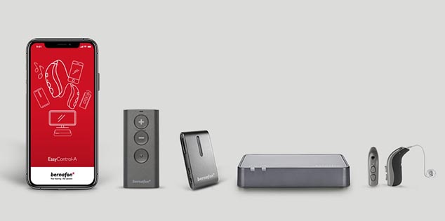 Bernafon accessories lined up, including Bernafon app on a smartphone, TV adapter, remote control, hearing aids and SoundClip-A